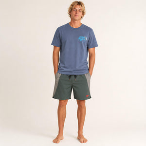 A man stands barefoot against a plain light background wearing a slate blue t-shirt with a light blue 'CHLAX' logo on the chest and dark green board shorts with gray side panels.