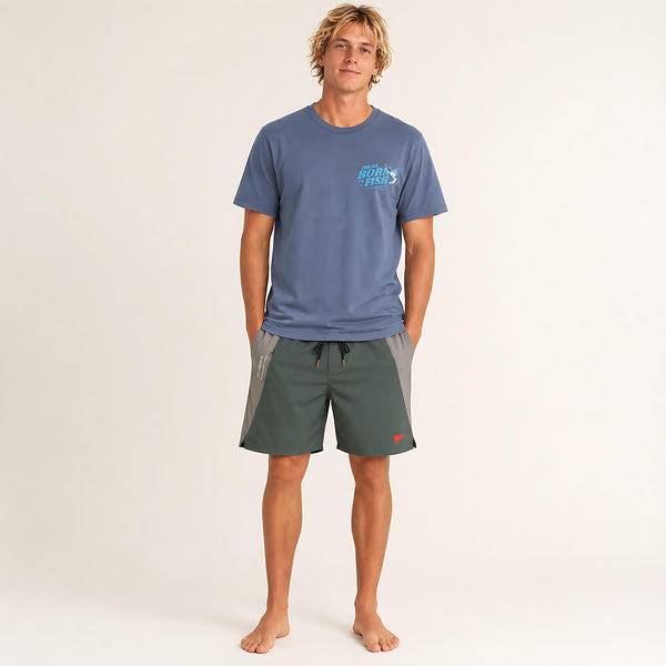 A man stands barefoot against a plain light background wearing a slate blue t-shirt with a light blue 'CHLAX' logo on the chest and dark green board shorts with gray side panels.