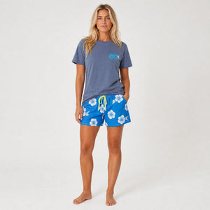 A woman modeling a slate blue t-shirt and bright blue floral swim shorts against a plain white studio background.