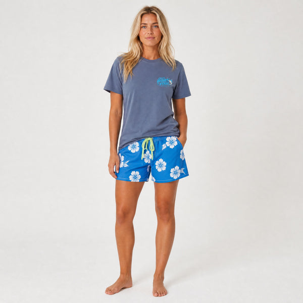 A woman modeling a slate blue t-shirt and bright blue floral swim shorts against a plain white studio background.