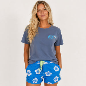 A woman modeling a slate blue t-shirt with a 'Great Barrier Reef' graphic on the left chest, paired with bright blue floral swim shorts, against a plain light gray background.