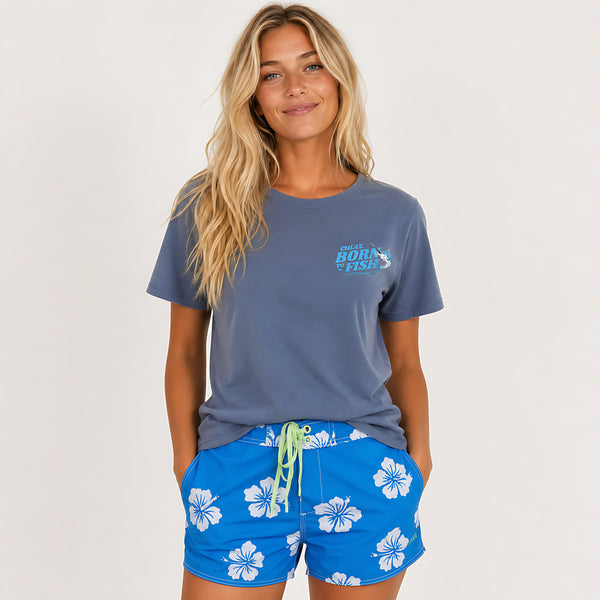 A woman modeling a slate blue t-shirt with a 'Great Barrier Reef' graphic on the left chest, paired with bright blue floral swim shorts, against a plain light gray background.