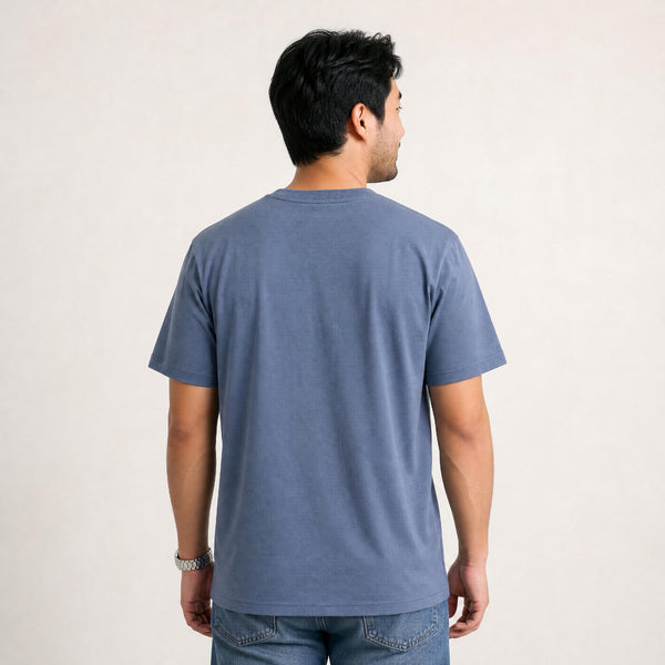 A man viewed from the back wearing a short-sleeve crewneck t-shirt in a muted slate blue color, paired with blue jeans against a plain off-white background.