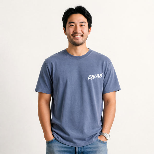 A man wearing a slate blue crewneck t-shirt with a white 'CHLAX' logo on the left chest, paired with blue jeans, standing against a plain off-white background.