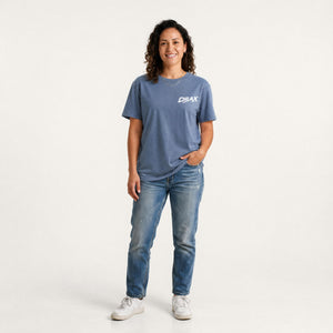 A woman modelling a slate blue crewneck t-shirt with a white 'CHLAX' logo on the chest, paired with blue jeans and white sneakers, against a plain white studio background.