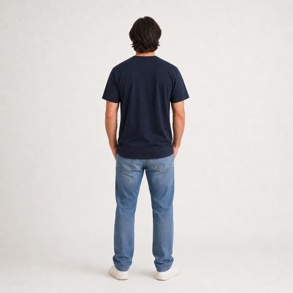 A man viewed from behind wearing a dark navy blue t-shirt, light blue jeans, and white sneakers, standing against a plain light gray studio background.