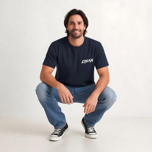 A man crouching in a studio setting, wearing a navy blue t-shirt with a white 'CHLAX' logo on the chest, blue jeans, and black canvas sneakers.