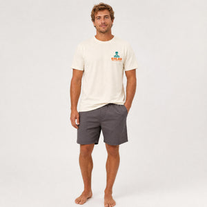 A man modeling a cream-colored short-sleeve t-shirt with a small green and orange 'CHLAX' logo on the left chest, paired with dark gray shorts, against a plain white studio background.