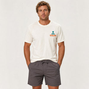A man wearing a cream-colored short-sleeve t-shirt with a small green and orange 'CHLAX' logo on the left chest, paired with dark gray drawstring shorts, standing against a light gray studio background.