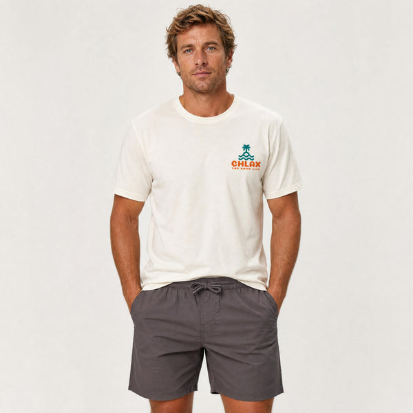 A man wearing a cream-colored short-sleeve t-shirt with a small green and orange 'CHLAX' logo on the left chest, paired with dark gray drawstring shorts, standing against a light gray studio background.