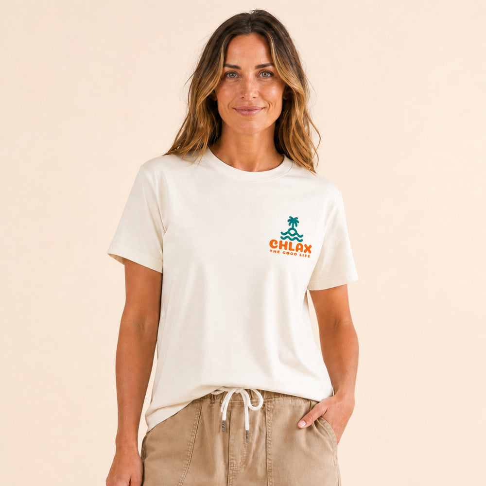 A woman wearing a cream-colored t-shirt with a colorful back graphic that reads 'AM PM CHLAX THE GOOD LIFE' with a palm tree and waves, paired with khaki shorts against a plain light background.