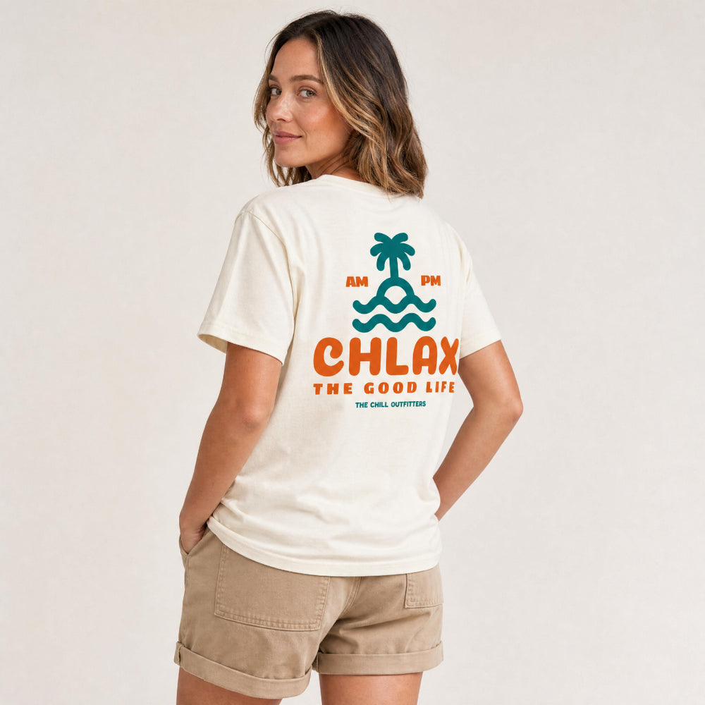 A woman wearing a cream-colored t-shirt with a colorful back graphic that reads 'AM PM CHLAX THE GOOD LIFE' with a palm tree and waves, paired with khaki shorts against a plain light background.