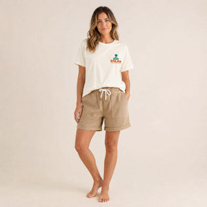 A woman modeling a cream-colored crewneck t-shirt with a small orange and green 'CHLAX' logo on the left chest, paired with beige drawstring shorts, against a solid light beige background.