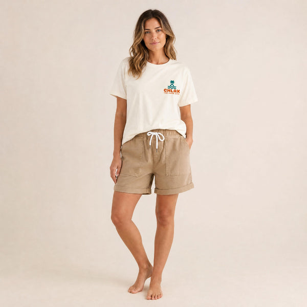 A woman modeling a cream-colored crewneck t-shirt with a small orange and green 'CHLAX' logo on the left chest, paired with beige drawstring shorts, against a solid light beige background.