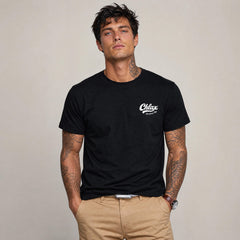 A man wearing a black crewneck t-shirt with a white 'Chlax' logo on the left chest, paired with khaki pants, standing against a neutral beige background.