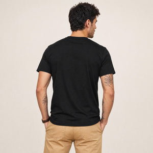 A man viewed from behind wearing a plain black short-sleeve t-shirt and tan pants, standing against a light beige studio background.
