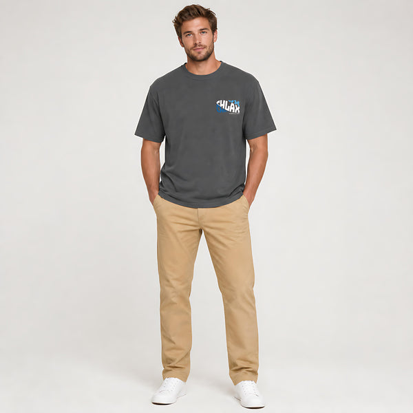A man modeling a dark gray crewneck t-shirt with a small blue and white graphic on the left chest, paired with beige pants and white sneakers, against a light gray studio background.