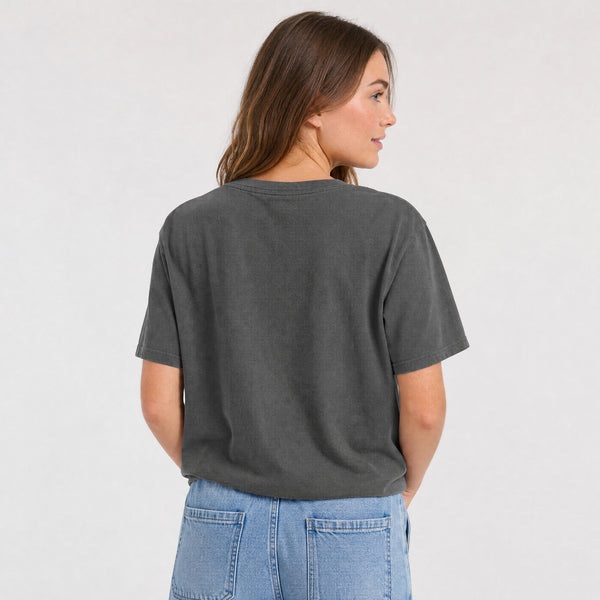A woman shown from the back wearing a dark charcoal gray short-sleeve t-shirt and light blue jeans, standing against a plain light gray studio background.