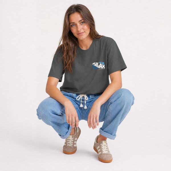 A woman crouching in a studio setting, wearing a dark charcoal gray t-shirt with a blue and white 'CHLAX' graphic on the left chest, paired with light blue jeans and beige sneakers.