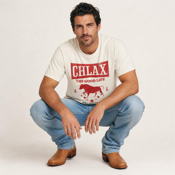 A man crouching in a studio setting wearing a cream-colored graphic t-shirt with a red bull design and the text 'CHLAX THE GOOD LIFE', paired with blue jeans and brown leather boots.