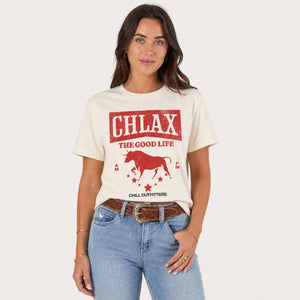 A woman modeling a cream-colored graphic t-shirt with a red bull design and the text 'CHLAX THE GOOD LIFE' and 'CHILL OUTFITTERS', paired with blue jeans and a brown belt, against a light beige background.