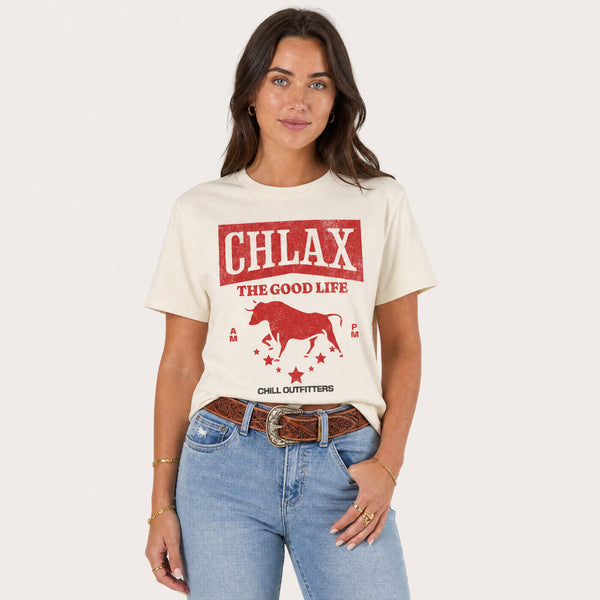 A woman modeling a cream-colored graphic t-shirt with a red bull design and the text 'CHLAX THE GOOD LIFE' and 'CHILL OUTFITTERS', paired with blue jeans and a brown belt, against a light beige background.