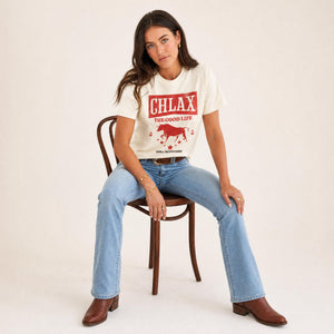 A woman sits on a wooden chair wearing a cream graphic t-shirt with a red 'CHLAX THE GOOD LIFE' logo and a horse illustration, paired with light blue jeans and brown leather boots, against a plain cream studio background.