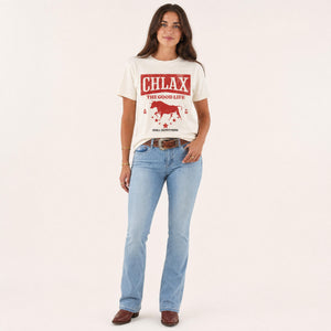 A woman modelling a cream-colored graphic t-shirt with a red western-style design that reads 'CHLAX THE GOOD LIFE' and 'CHILL OUTFITTERS', paired with light-wash blue jeans and brown shoes, against a solid off-white background.