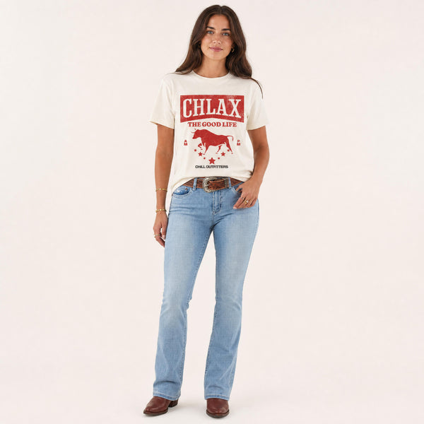 A woman modelling a cream-colored graphic t-shirt with a red western-style design that reads 'CHLAX THE GOOD LIFE' and 'CHILL OUTFITTERS', paired with light-wash blue jeans and brown shoes, against a solid off-white background.