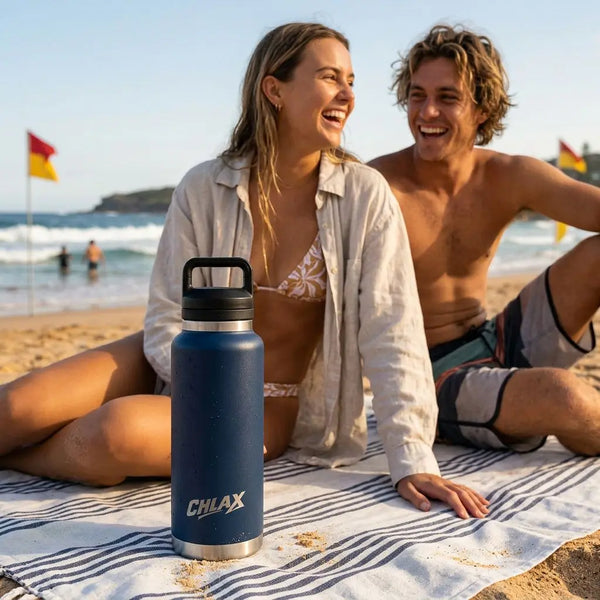 A navy blue insulated water bottle with a black handle and silver CHLAX logo sits on a striped beach towel on a sandy beach, with a smiling couple in swimwear sitting behind it.