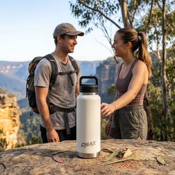 A white insulated water bottle with a black lid stands on a rock outdoors, with a man and woman hiking in the background. The bottle has the brand name CHILAX printed on the front. #driftwood