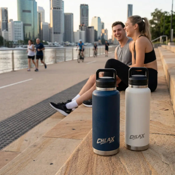 Two insulated water bottles from CHLAX, one navy blue and one white, sitting on a stone walkway next to a waterfront with a city skyline in the background. A smiling man and woman are seated nearby on the stone ledge.