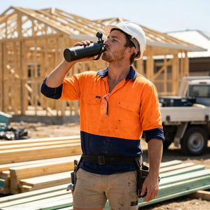 A man on a construction site wearing a high-visibility orange and navy work shirt, khaki shorts, and a white hard hat, drinking from a black insulated water bottle.
