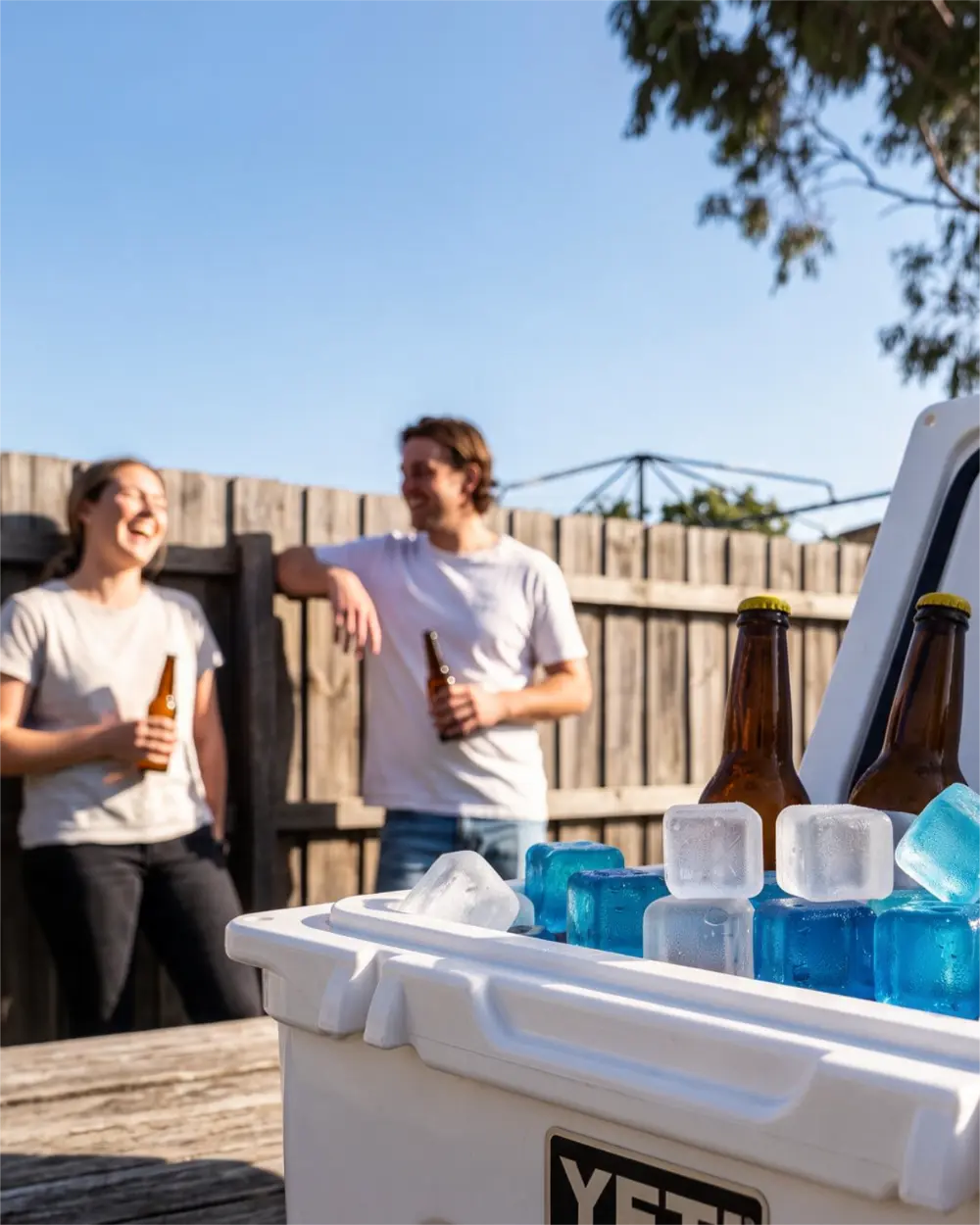 A white YETI cooler in the foreground with blue reusable ice cubes and brown beer bottles, with two people standing and laughing in a sunny backyard behind a wooden fence.
