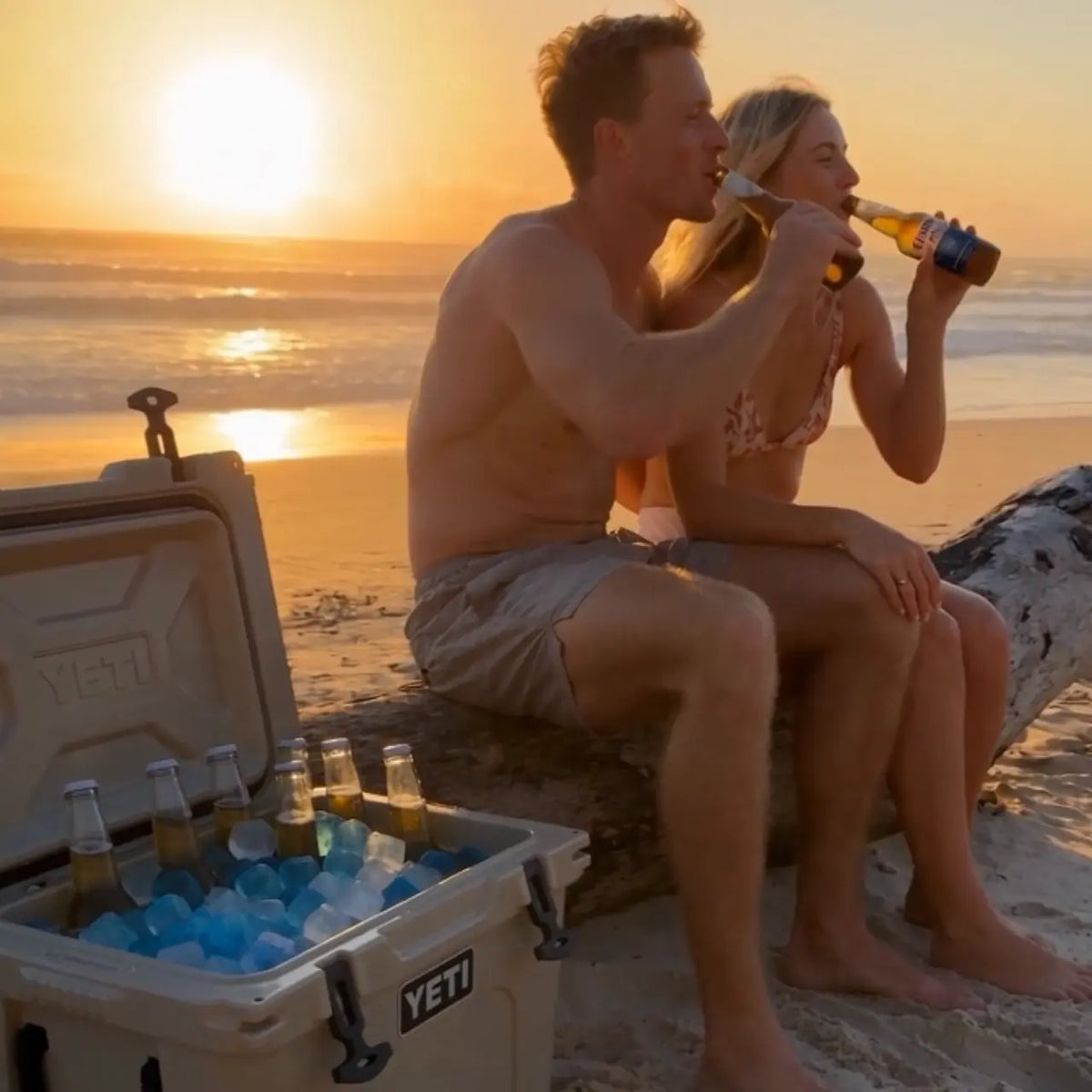 A man and woman sit on a beach log at sunset while drinking beer. A YETI cooler filled with ice and beer bottles sits in the foreground on the sand.