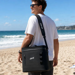 A man wearing sunglasses and a white t-shirt stands on a sandy beach carrying a black CHLAX cooler bag with a shoulder strap, with the ocean and blue sky in the background.