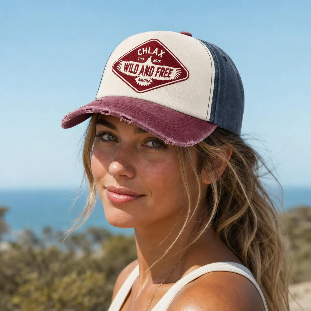 A woman outdoors wearing a distressed trucker cap with a cream front panel, dark blue side panels, and a maroon brim. The front patch reads 'CHLAX Wild and Free'.