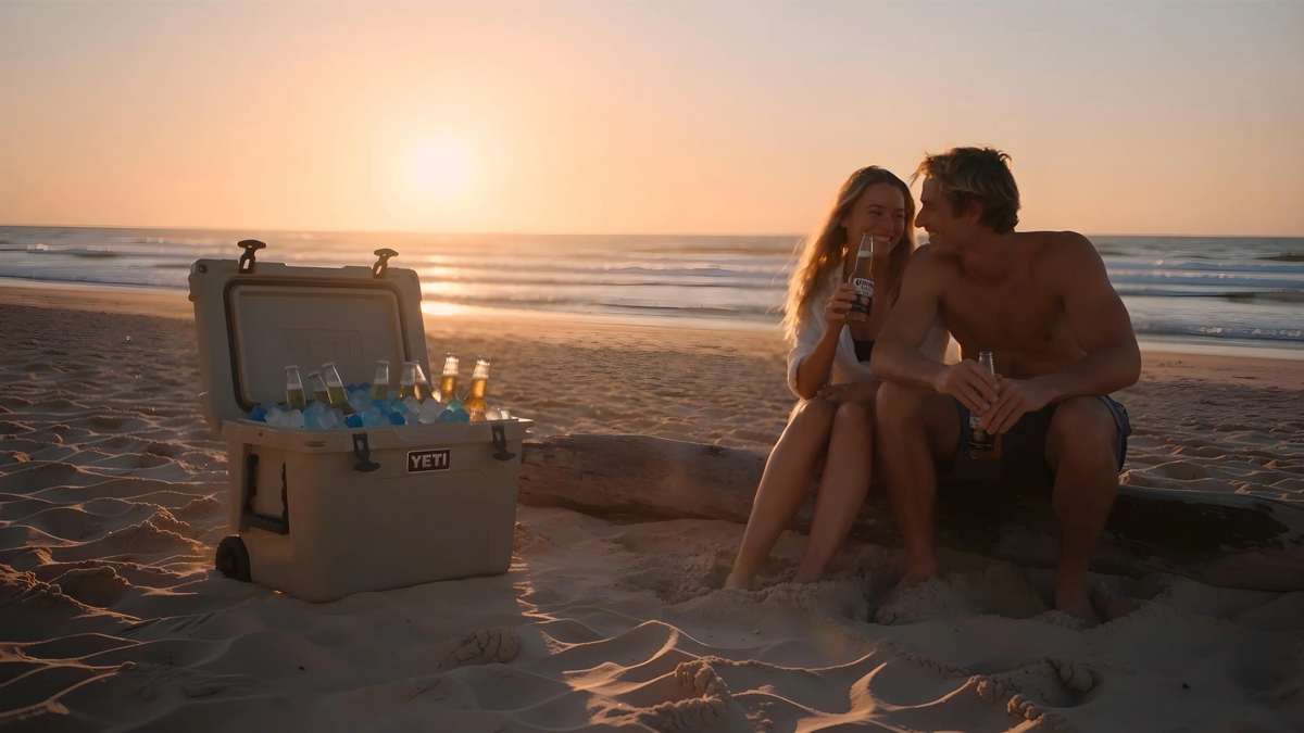 A YETI cooler filled with beer bottles sits open on a sandy beach at sunset, next to a couple sitting on a log and holding drinks.