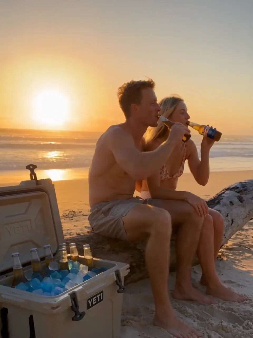 A man and a woman sit on a log on a beach at sunset, drinking from bottles. A YETI cooler filled with ice and bottles sits on the sand in the foreground.