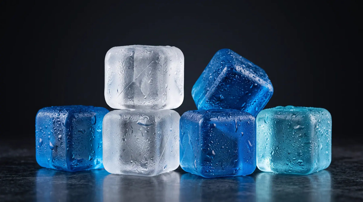 Six translucent reusable ice cubes in shades of blue, cyan, and clear, arranged on a dark reflective surface against a black background.