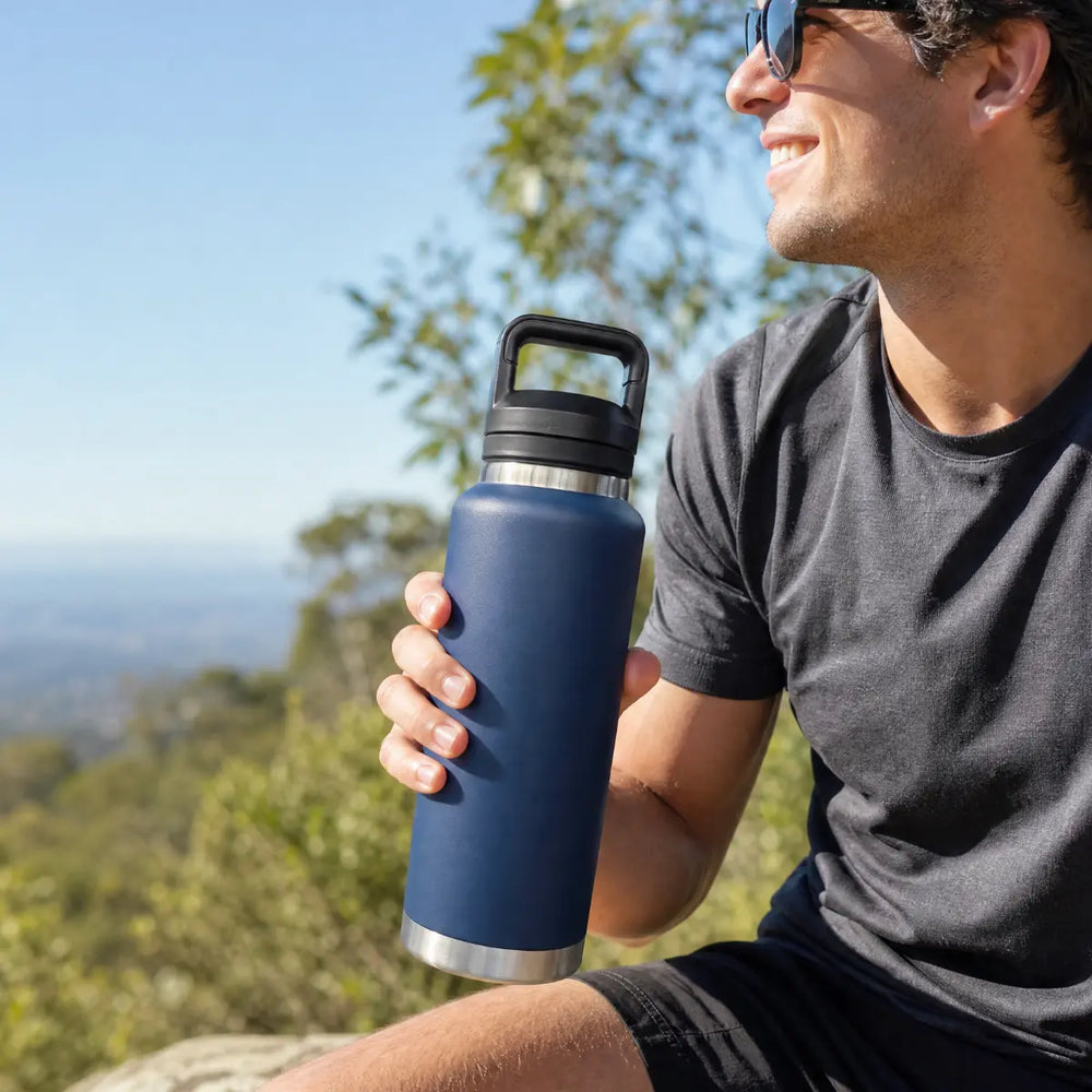 A man wearing sunglasses and a dark gray t-shirt holds a navy blue insulated water bottle with a black lid and silver base, set against a blurred outdoor background of trees and sky.