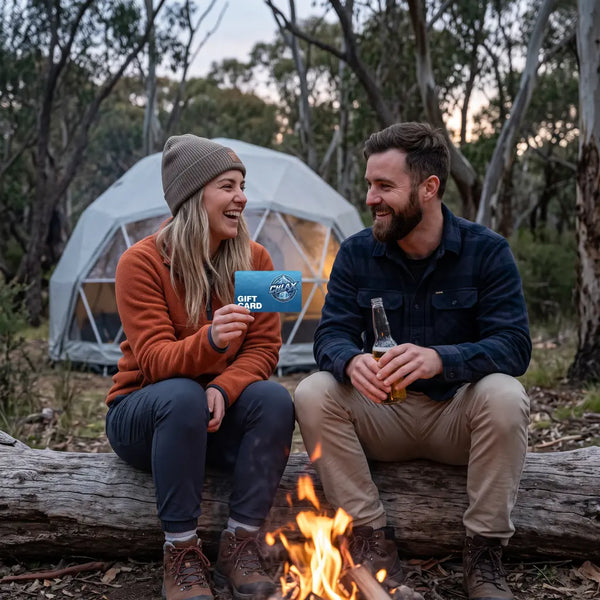 A man and a woman sit on a log by a campfire in a wooded outdoor setting, with a geodesic-style camping tent behind them. The woman holds a blue card, and the man holds a glass bottle.