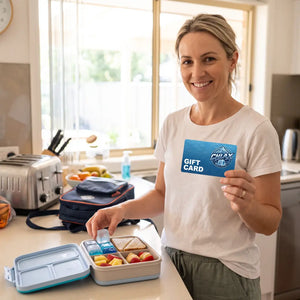 A woman in a white t-shirt stands in a bright kitchen holding a blue Galaxy gift card next to an open bento lunch box with fruit and sandwich halves.