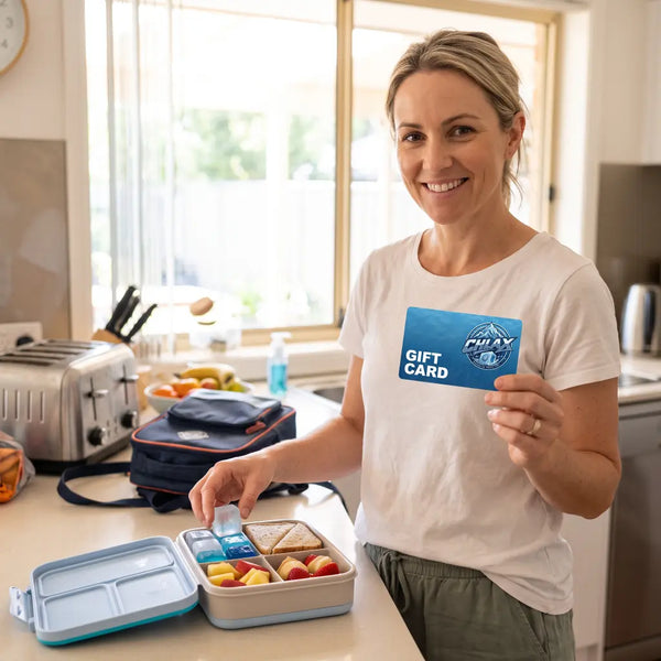 A woman in a white t-shirt stands in a bright kitchen holding a blue Galaxy gift card next to an open bento lunch box with fruit and sandwich halves.