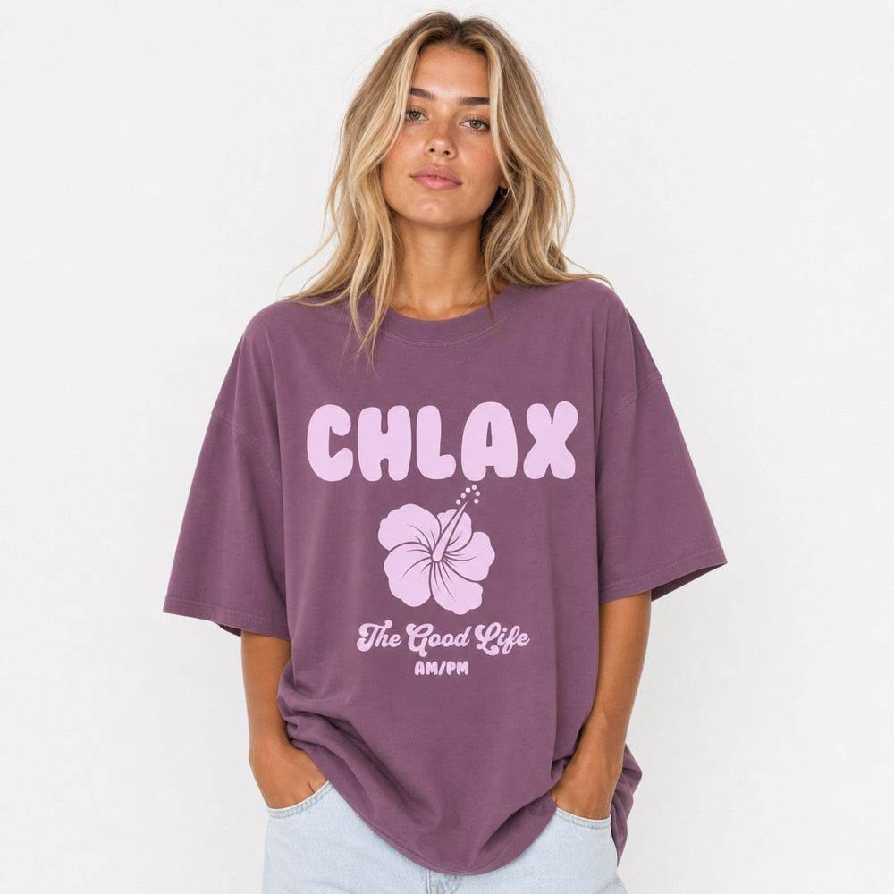 A woman wearing an oversized mauve purple t-shirt with a light pink graphic that reads 'CHLAX' and 'The Good Life AM/PM' with a hibiscus flower, paired with light blue jeans against a white studio background.