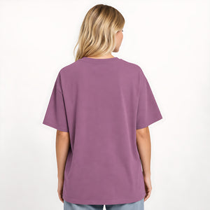 A woman viewed from behind wearing an oversized short-sleeve t-shirt in a dusty purple color, paired with light blue jeans, against a plain white background.