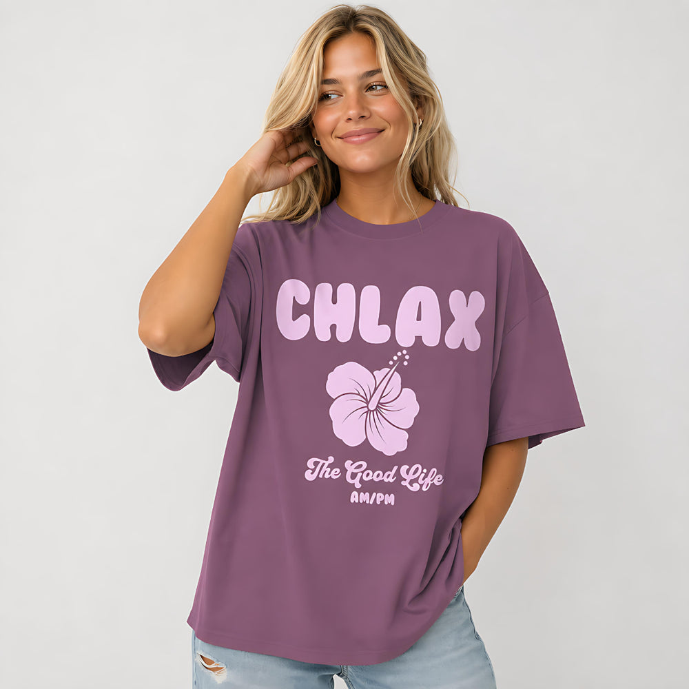 A woman wearing an oversized mauve purple t-shirt with a light pink graphic that reads 'CHLAX' and 'The Good Life AM/PM' with a hibiscus flower, paired with light blue jeans against a white studio background.