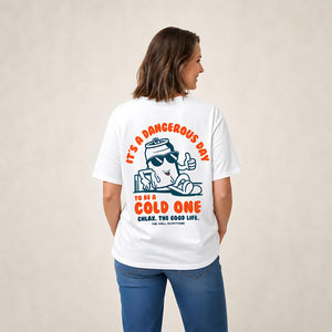 A woman shown from the back wearing a white graphic t-shirt with a cartoon character and orange text that reads 'IT'S A DANGEROUS DAY TO BE A COLD ONE' and 'CHLAX. THE GOOD LIFE.