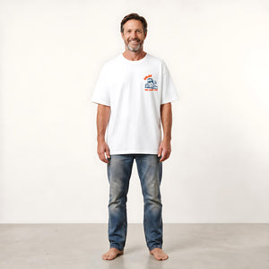 A man wearing a white crewneck t-shirt with a small red and blue graphic on the left chest, paired with blue jeans, standing against a plain off-white studio background.