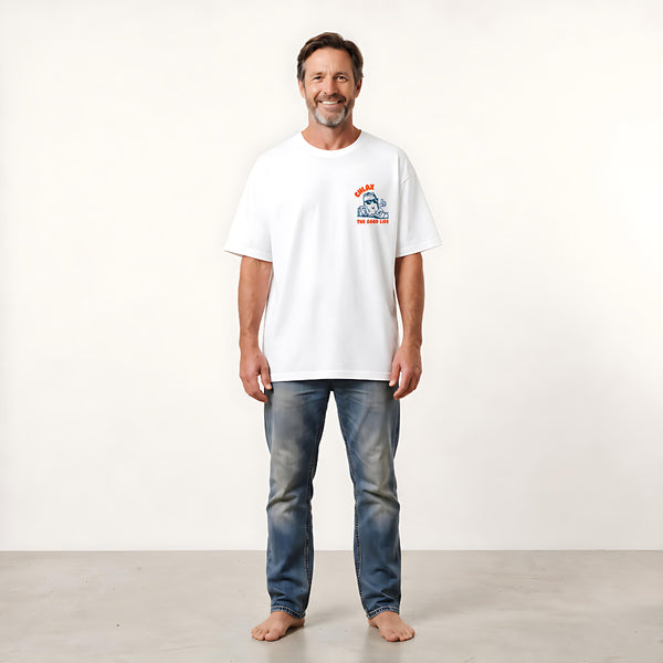 A man wearing a white crewneck t-shirt with a small red and blue graphic on the left chest, paired with blue jeans, standing against a plain off-white studio background.
